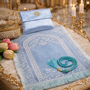 May include: A light blue prayer rug with a floral design, a matching satin pouch with a gold embellishment, and a turquoise prayer bead set. The rug has a fringed edge and is displayed with candles and decorative items.