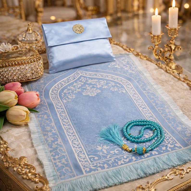 May include: A light blue prayer rug with a floral design, a matching satin pouch with a gold embellishment, and a turquoise prayer bead set. The rug has a fringed edge and is displayed with candles and decorative items.