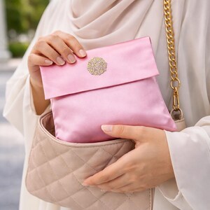 May include: A pink satin clutch with a gold-colored decorative embellishment. The clutch is held inside a quilted beige handbag with a gold chain strap. The image showcases a coordinated set of accessories.