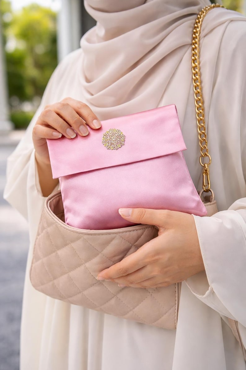 May include: A pink satin clutch with a gold-colored decorative embellishment. The clutch is held inside a quilted beige handbag with a gold chain strap. The image showcases a coordinated set of accessories.