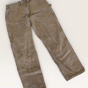 Puede incluir: Pantalones de trabajo marrones con rodillas reforzadas y múltiples bolsillos. Los pantalones tienen un diseño clásico de ropa de trabajo, con cierre de botón y cremallera, trabillas para el cinturón y detalles de remaches. La tela duradera sugiere un uso rudo.