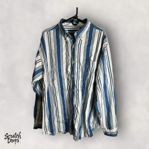 Può includere: Camicia a maniche lunghe a righe verticali blu e bianche, appesa a una gruccia nera. La camicia ha una tasca sul petto e il colletto riporta la scritta "Field & Stream".