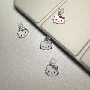 May include: Four white Hello Kitty charms with different colored bows. The charms are attached to clear plastic strings. The charms are on a white surface.