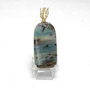Könnte beinhalten: Ein Anhänger mit einem hellblauen, braunen und weißen Opal-Stein mit einer goldenen Drahtummantelung. Der Stein ist rechteckig und hat eine glatte, polierte Oberfläche.