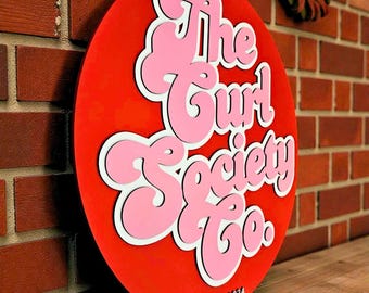May include: A round, red sign with the text "The Curl Society Co." in pink, outlined in white. The sign is leaning against a brick wall. The text "EST. 2024" is at the bottom. The sign is a decorative item.