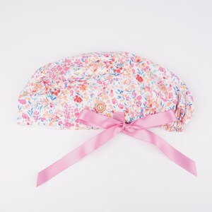 Puede incluir: Un gorro quir&uacute;rgico blanco con estampado floral rosa y un lazo de cinta de sat&eacute;n rosa. El gorro tiene un cierre de bot&oacute;n.