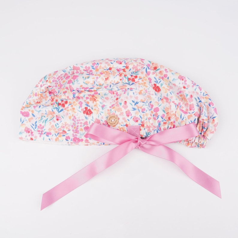 Puede incluir: Un gorro quir&uacute;rgico blanco con estampado floral rosa y un lazo de cinta de sat&eacute;n rosa. El gorro tiene un cierre de bot&oacute;n.