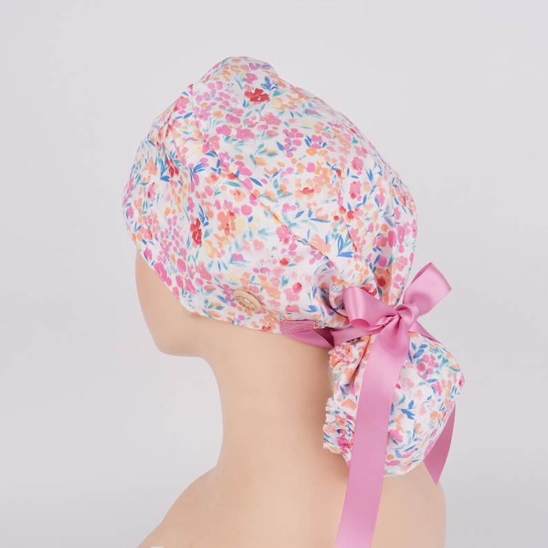 Puede incluir: Un gorro de quir&oacute;fano blanco con estampado floral rosa y un lazo rosa. El gorro tiene un bot&oacute;n en la parte superior.