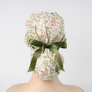 May include: A patterned scrub cap in white and green, decorated with squirrels, leaves, and carrots. It features a green satin bow and a button closure. This cap is designed for medical professionals and is a stylish accessory.