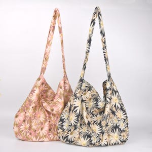 May include: Two floral print tote bags. One bag is a light pink with white and yellow daisy-like flowers. The other bag is black with white and yellow daisy-like flowers. Both bags have long straps and are made of a soft fabric.