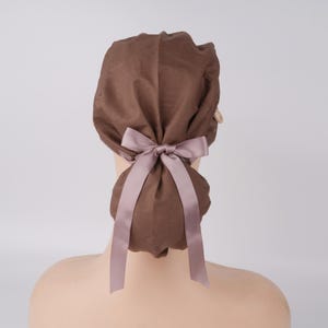 May include: A brown fabric hair covering with a light purple bow and long ribbons. The hair accessory is tied at the back of the head, with the bow centered. The fabric appears to be soft and slightly textured.
