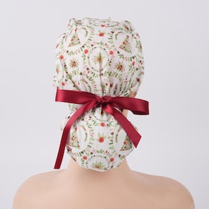 May include: A white scrub cap with a festive Christmas tree and floral pattern. The cap is tied with a burgundy ribbon bow at the back. The cap is designed for medical professionals.