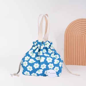 May include: A blue drawstring bag with a white floral pattern and light green centers. The bag has a cream-colored handle and a drawstring closure. A small, round tag with text is attached to the bag.