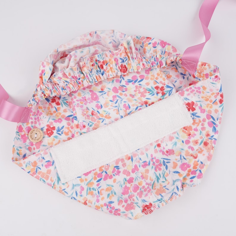 Puede incluir: Un portabeb&eacute;s blanco con estampado floral rosa y un inserto acolchado blanco. El portabeb&eacute;s tiene correas rosas y un cierre de bot&oacute;n.