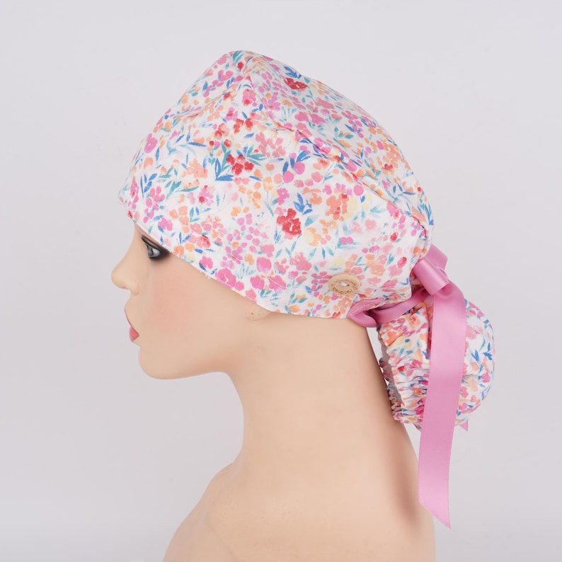 Puede incluir: Un gorro de quir&oacute;fano blanco con estampado floral en tonos rosa, naranja y azul. El gorro tiene un lazo de cinta rosa y un bot&oacute;n en el lateral.