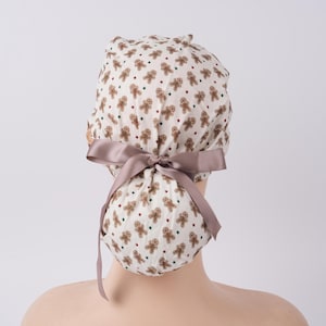 May include: A white and tan patterned scrub cap featuring a repeating gingerbread man design with red, green, and blue star accents. The cap is tied with a taupe satin ribbon. This festive cap is perfect for healthcare workers.