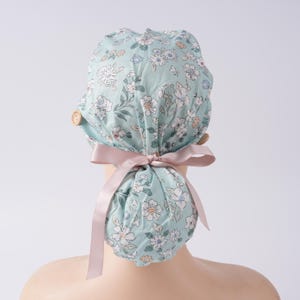 May include: A light blue floral patterned scrub cap with a pink ribbon tied in a bow. The cap features a repeating pattern of white and yellow flowers and green leaves. Two wooden buttons are visible on the side of the cap.