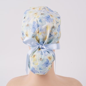 May include: A floral patterned scrub cap featuring a light blue ribbon bow. The cap is adorned with a watercolor design of blue and yellow flowers on a white background. The cap is designed to be tied at the back.