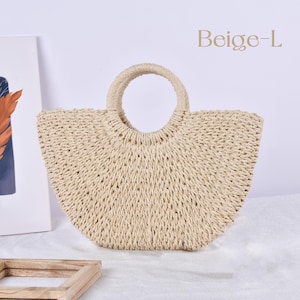 Op de afbeelding: Een beige geweven strooien draagtas met een ronde handgreep. De tas heeft een afgeronde vorm en is gemaakt van natuurlijk gekleurd stro. De tekst "Beige-L" staat in de rechterbovenhoek. De tas staat op een wit oppervlak.