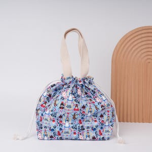 May include: A light blue drawstring bag with a cream-colored handle and drawstring. The bag is patterned with illustrations of characters from Alice in Wonderland in red, white, and brown. The bag is approximately 8 inches tall and 7 inches wide.