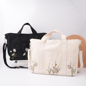 Può includere: Due borse tote in tela, una nera e una bianco sporco, ciascuna con ricami di margherite. La borsa nera ha una tracolla e la borsa bianco sporco ha una tracolla e due manici. La parola "Flowers" è visibile sulla borsa nera.