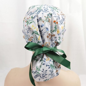 May include: A white scrub cap with a floral print and green ribbon ties. The cap is tied at the back of the head.