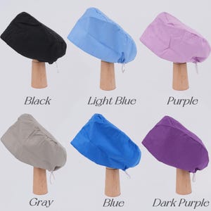 Op de afbeelding: Een verzameling van zes chirurgische mutsen in verschillende kleuren, waaronder zwart, lichtblauw, paars, grijs, blauw en donkerpaars. Elke muts wordt getoond op een houten standaard, met de kleurnaam eronder.