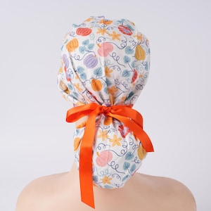 May include: A white scrub cap with a colorful pumpkin pattern in orange, pink, purple, and yellow. The cap is tied with a bright orange ribbon. The design includes vines and flowers.