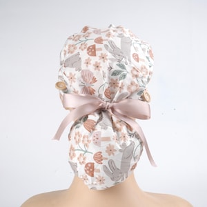 May include: A white scrub cap with a floral and bunny print in shades of brown, peach, and gray. The cap features a light pink satin ribbon and two wooden buttons. The cap is designed to be tied at the back.