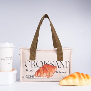 Peut inclure: Un sac à déjeuner en toile beige avec des sangles brunes. Le sac a un graphique d'un croissant avec le texte "CROISSANT" et "Pain" et "Délicieux".