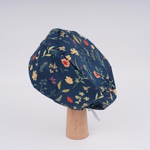 May include: A navy blue surgical scrub cap with a floral pattern in yellow, red, and purple. The cap is displayed on a wooden stand and has a white tie in the back. The floral design includes various flowers and leaves.