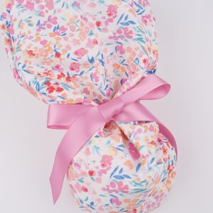 Puede incluir: Un gorro de quir&oacute;fano blanco con estampado floral rosa y una cinta de raso rosa atada alrededor.