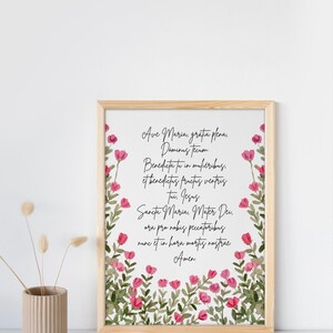 May include: A framed print with a white background and pink floral border. The print features the text "Ave Maria, gratia plena, Dominus tecum Benedicta tu in mulieribus, et benedictus fructus ventris tui. Jesus Sancta Maria, Mater Dei, ora pro nobis peccatoribus nunc et in hora mortis nostrae Amen."