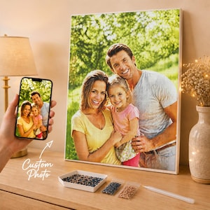 May include: A custom diamond painting kit featuring a family portrait of a man, woman, and child in a park. The set includes a framed canvas, a smartphone displaying the original photo, a small white tray with colorful beads, and a stylus tool.
