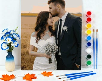May include: A paint-by-numbers kit featuring a couple in wedding attire, with a field backdrop. The kit includes paint pots in various colors, paintbrushes, and a vase of blue and white flowers. Orange leaves are scattered on the white surface.