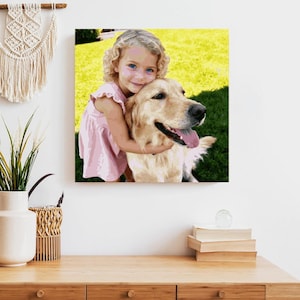 Op de afbeelding: Een vierkante canvas print met een jong meisje met blond krullend haar dat een golden retriever omhelst. Het meisje draagt een roze jurk. De achtergrond toont groen gras en een gele lucht.