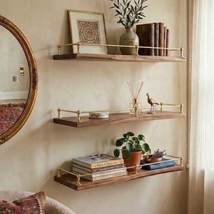 Puede incluir: Tres estantes flotantes de madera con barandillas de latón están montados en una pared beige. Los estantes exhiben objetos decorativos como libros, plantas, una imagen enmarcada, un jarrón con vegetación y una pequeña figura de pájaro. Un espejo con marco dorado es visible a la izquierda.