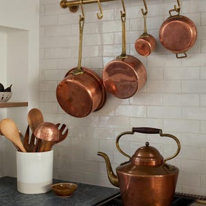 May include: A kitchen scene featuring copper cookware and a copper kettle. The pans hang from a brass rail, and the kettle sits on a hob. Wooden utensils are stored in a white and beige ceramic container.