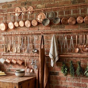 Könnte beinhalten: Eine Küche mit Kupfer-Kochgeschirr und Utensilien, die an Kupferstangen an einer Backsteinmauer hängen. Holzlöffel, Schneebesen und Schöpflöffel sind ausgestellt. Eine Schürze und Kräuter ergänzen die rustikale Einrichtung.