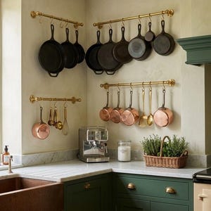 May include: A kitchen scene featuring cast iron skillets and copper pots hanging from brass pot racks. The kitchen has green cabinets, a marble countertop, and a copper sink. A basket of herbs sits on the counter.