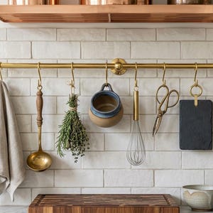 Puede incluir: Una cocina con una barra de latón para colgar utensilios y un paño de cocina. Incluye un cucharón, hierbas, un cuenco pequeño, un batidor, tijeras, una pizarra y un cuchillo. El fondo es de azulejos blancos. La barra mide 60 cm de largo.