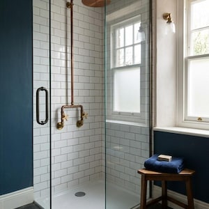 May include: A contemporary shower room with white brick-effect tiles and a glass screen. Copper shower fittings, including a rain shower head and taps, are visible. A small wooden stool holds a blue towel and soap near a window.