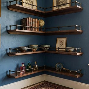 May include: Dark wood corner shelves with metal railings are mounted on a blue wall. The shelves hold books, bowls, framed art, and decorative bottles. The shelves are arranged in a corner, maximizing space and adding a touch of elegance.