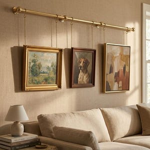 May include: Three framed artworks hanging from a gold-colored rod. The art includes a landscape painting, a dog portrait, and an abstract piece. The frames are various colors, including gold and brown. The rod is suspended by chains.