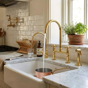 Op de afbeelding: Een gootsteen met een gouden kraan en sproeier. Water stroomt in een koperen pan. Een houten afdruiprek, glas en zeepdispenser staan op het marmeren aanrecht. Een plant in een pot staat op de vensterbank.