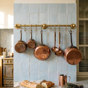 May include: A kitchen scene featuring copper cookware hanging on a brass rack against a light blue tiled wall. A loaf of bread, butter, and a copper mug sit on a marble countertop, creating a rustic, inviting atmosphere.