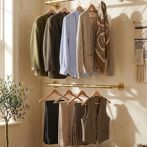 May include: A display of clothing on brass rods, including jackets, a striped shirt, a cardigan, a patterned scarf, trousers, and a vest. The items are on wooden hangers. A small potted olive tree and a vintage trunk are in the foreground.