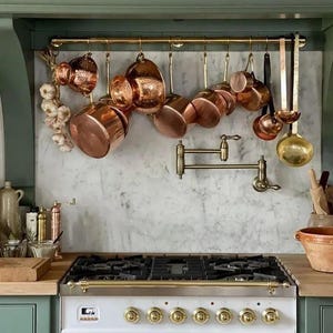 May include: A kitchen scene featuring copper cookware hanging above a white stove with brass accents. The copper pots and pans are suspended from a brass rack against a marble backsplash. A bundle of garlic hangs nearby.