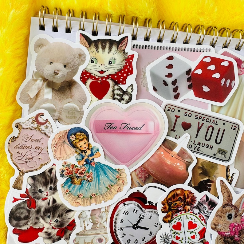 May include: A collection of stickers featuring a beige teddy bear, a kitten with a red bow, two dice with hearts, a Too Faced heart-shaped compact, a girl with an umbrella, a candle, a heart-shaped clock, and a rabbit on a spiral-bound notebook.
