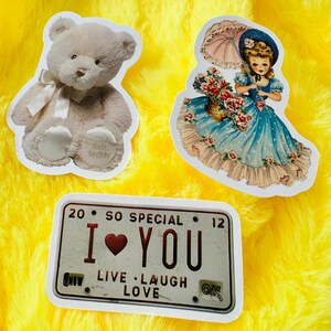 May include: Three stickers featuring a light beige teddy bear with a white ribbon, a girl in a blue dress holding a pink umbrella and a basket of flowers, and a rectangular license plate graphic with the text 20 SO SPECIAL 12 I LOVE YOU.